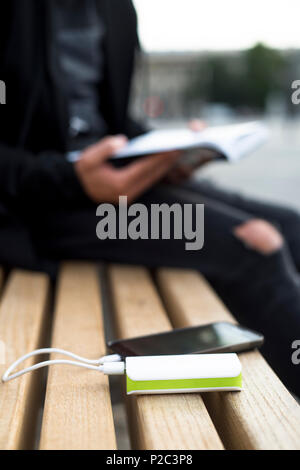 Ein junger kaukasischer Mann ein Buch lesen, in einer Bank sitzen auf der Straße, während sein Smartphone ist charching zu einem powerbank im Vordergrund gesteckt Stockfoto