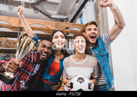 Aufgeregt jungen multiethnischen Freunde mit Fußball und Trophäe zusammen jubeln zu Hause Stockfoto