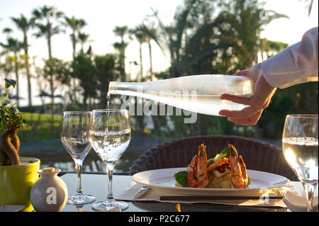 Ort Einstellung mit Wein Gläser, Riesengarnelen und Hand des Kellners Wasser im Garten Restaurant mit Palmen im BAC-Dienst Stockfoto