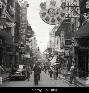 Die 1950er Jahre, historisch, ein Bild von J Allan Cash aus dieser Zeit, das eine Straße in der Altstadt in Hongkong, Asien, hinunterblickt und Menschen zeigt, die auf der Straße gehen, Geschäfte, chinesische Schilder und die Architektur der Gegend, mit den Wohnhäusern über den Einzelhandelsgeschäften. Auch auf dem Bild, geparkt auf der Straße, ein Auto der Ära mit einem Nummernschild, 423. Stockfoto