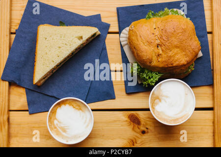 Zwei Becher mit Kaffee auf einer hölzernen Oberfläche. In der Nähe liegen ein Sandwich und einen Burger. Fast Food Stockfoto