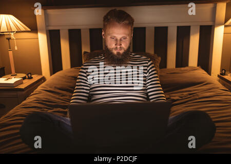 Mann sitzt auf Bett im dunklen Zimmer mit seinem Laptop Stockfoto