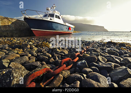 Boot vertäut am Steg mit Kiesstrand und roten rostigen Kette in Richtung der Küste im Vordergrund führende Stockfoto