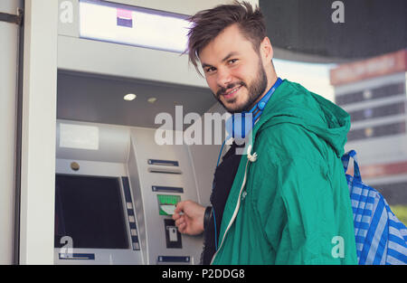 Junge hipster Kerl in Kopfhörer bei Kamera Lächeln beim Einsetzen der Kreditkarte in Geldautomaten Bargeld zu erhalten Stockfoto