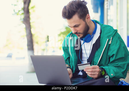 Stilvolle hipster Mann sitzt auf der Straße mit Laptop auf den Knien und das Tun von on-line-Kauf mit Kreditkarte Stockfoto