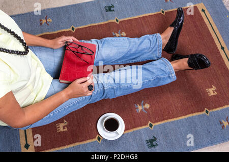 In der Nähe der jungen Frau legere Kleidung und Kette sitzen auf Teppich, rote vintage Buch, eine Tasse Kaffee, und schwarze Brille. Stockfoto