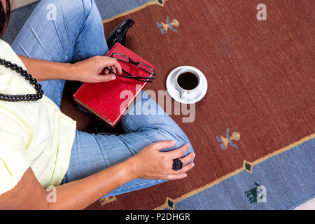 In der Nähe der jungen Frau legere Kleidung und Kette sitzen auf Teppich, rote vintage Buch, eine Tasse Kaffee, und schwarze Brille. Stockfoto