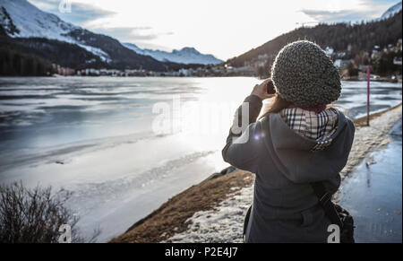 Frau nimmt ein Bild von einem zugefrorenen See mit einer Kamera bei Sonnenuntergang in St. Moritz Stockfoto