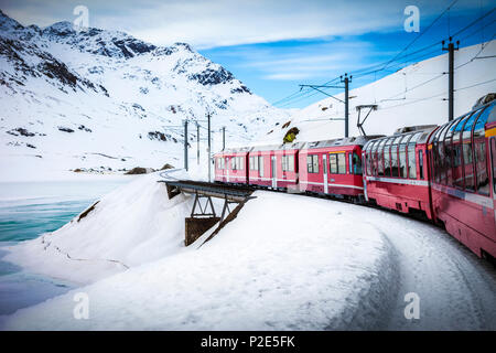 Bernina Express Zug, einer der höchsten Bahn der Welt, geht über eine kleine Brücke im verschneiten Berg in der Nähe eines zugefrorenen See Stockfoto