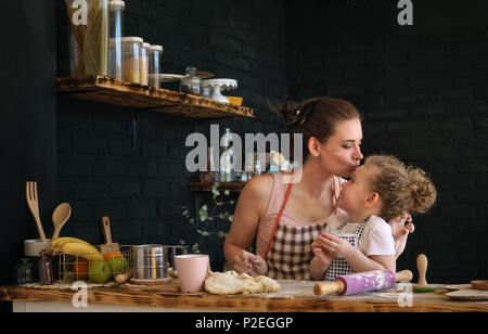 Junge Mutter und Tochter bereiten Sie Cookies in der Küche. Sie sind in den Schürzen. Frau küsse Kind auf die Stirn. Zeit mit der Familie. Stockfoto