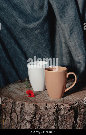 Neue Tassen auf einer hölzernen Stump, mit einer roten Blume, auf grauem Stoff Hintergrund. Immer noch leben. foto Utensilien Stockfoto