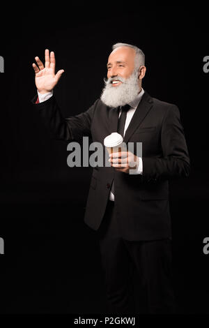 Halbe Länge geschossen von lächelnden Geschäftsmann im Anzug mit einer Tasse Kaffee und winkte mit der Hand auf jemanden Stockfoto