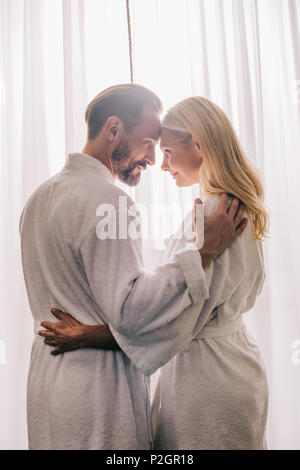 Seitenansicht der glücklichen wunderschöne reife Paar in Liebe tragen Bademäntel und lächeln einander im Hotel Zimmer Stockfoto