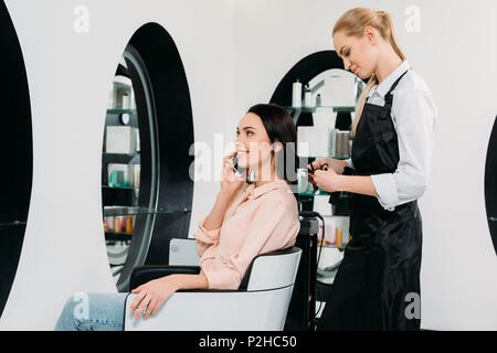 Frau sprechen von Smartphone, während Friseur Haare schneiden Stockfoto