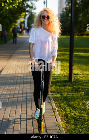 Foto des lockigen Haaren athletische Frau reiten Roller im Park im Sommer Tag Stockfoto