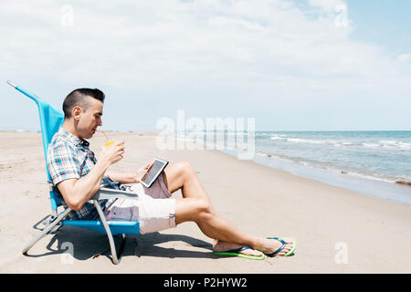 Nahaufnahme eines jungen kaukasischen Mann, sitzend auf einem Liegestuhl am Strand, mit einem digitalen Tablet beim trinken einen erfrischenden Drink orange Stockfoto