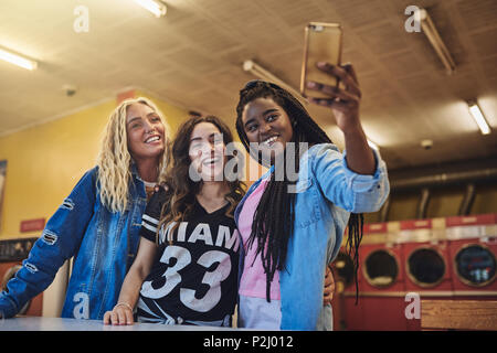 Lächelnde junge weibliche Freunde in einem Waschsalon stehen unter selfies während Wäsche zusammen tun Stockfoto