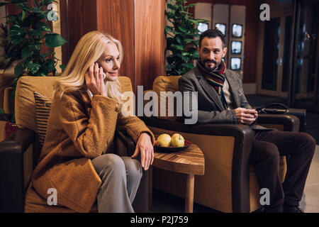 Paar mittleren Alters mit Smartphones beim Sitzen und Warten im Hotel Flur Stockfoto