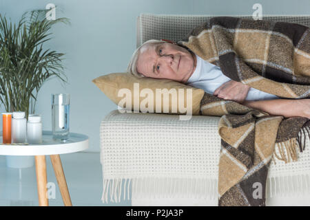 Senior kranken Mann in Plaid liegt auf einem Sofa im Zimmer mit Medikationen auf Tabelle Stockfoto