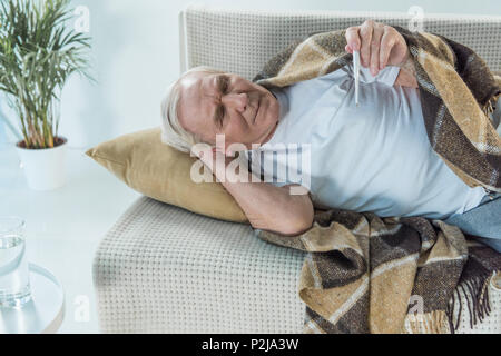 Senior kranken Mann in Plaid liegt auf dem Sofa und Prüfungen abgedeckt Thermometer Stockfoto