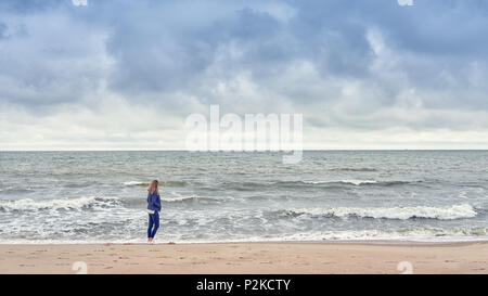 Frau zu Fuß entlang der Kante der Brandung am Strand in einer blauen Jeans Outfit mit Blick aufs Meer in einer kalten, bewölkten Tag mit Platz zum Kopieren Stockfoto