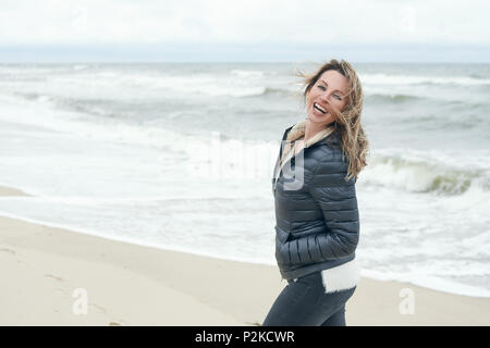Vivacious lacht unbeschwert Frau auf einem windgepeitschten Strand mit ihr langes Haar weht im Wind und rauer See, mit Kopie Raum Stockfoto