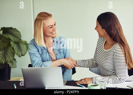 Zwei lächelnde junge Unternehmerinnen an einem Tisch sitzen die Hände schütteln zusammen, während in einem Büro arbeiten Stockfoto