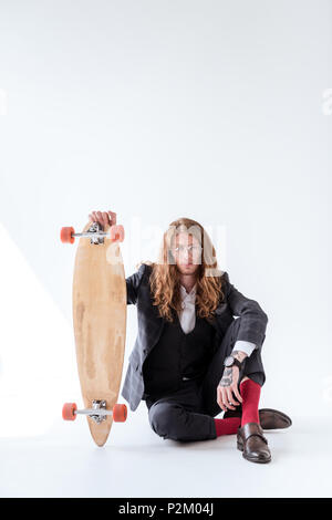 Stilvolle Geschäftsmann mit lockigem Haar saß auf dem Boden und halten Skateboard Stockfoto