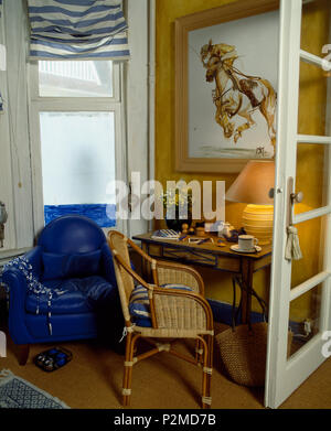 Großes Gemälde an der Wand über dem korbstuhl und einem kleinen Tisch im Wohnzimmer mit gestreiften Vorhang am Fenster oben Blau Leder Sessel Stockfoto