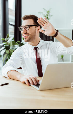 Lächelnd Geschäftsmann mit Laptop am Arbeitsplatz arbeiten und winken zu jemand Stockfoto