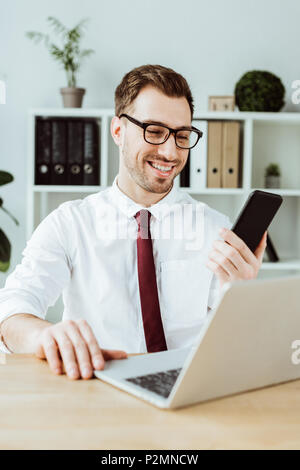 Gut aussehender Geschäftsmann mit Smartphone und Laptop am Arbeitsplatz in modernen Büro Stockfoto