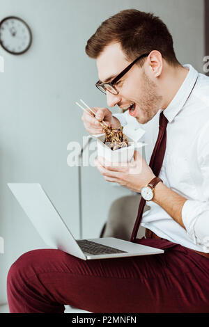 Stilvolle Geschäftsmann Essen Nudeln beim Arbeiten mit Laptop im Büro Stockfoto