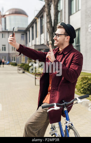 Stilvolle lächelnden Mann anzeigen Sieg unterzeichnen und unter selfie auf dem Smartphone während lehnte sich auf dem Fahrrad Stockfoto