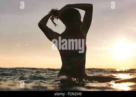 Silhouette einer Frau nachstellen Haar beim Sitzen auf Surfen im Ozean auf Sonnenuntergang Stockfoto