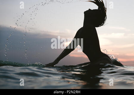 Silhouette einer Frau treibend Haar, während er auf das Surfen im Meer bei Sonnenuntergang Stockfoto