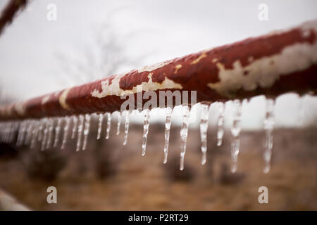 Eiszapfen hängen von einem rostigen alten Rohr oder Pfosten in einem Hof oder Zaun an einem eiskalten Wintertag in der Landschaft Stockfoto