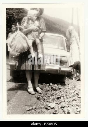 Die tschechoslowakische SOZIALISTISCHE REPUBLIK - ca. 1980 s: Vintage Foto zeigt Mutter hält ein kleines Mädchen. Sie stehen vor dem Auto (MARKE LADA), Auto in der UDSSR gemacht. Retro Schwarz/Weiß-Fotografie. Stockfoto