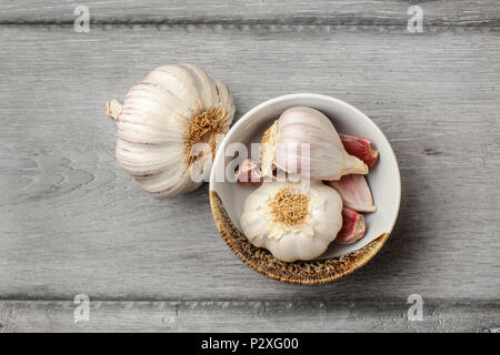 Table Top Blick auf Knoblauch Zwiebeln und geschälten Knoblauchzehen in kleine keramische Schüssel auf grau Holz Schreibtisch platziert. Stockfoto