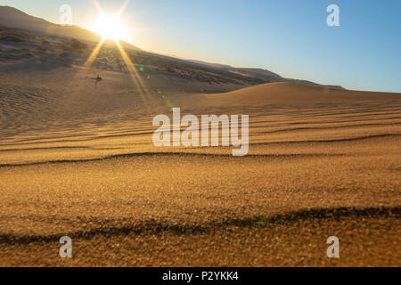 Sun steigt über Sossusvlei Dünen casting lens flare über Landschaft aus Sand und Wind geblasen Welligkeit Muster Stockfoto