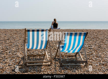 Zwei Liegestühle am Strand von Brighton mit einer jungen Frau sitzt in der Sonne. Stockfoto