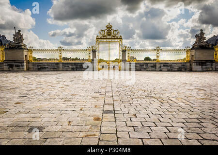 Gold Tor und Zaun am Eingang des Chateau de Versailles, mit gepflasterten Innenhof, gegen einen dramatischen, stark bewölkt, blauer Himmel. Stockfoto