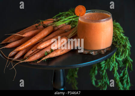 Vegetarische und diätetische Konzept mit einem Glas Karottensaft und einen Haufen frisch geerntete Möhren, auf einem schwarzen runden Tisch, bei schwachem Licht. Stockfoto