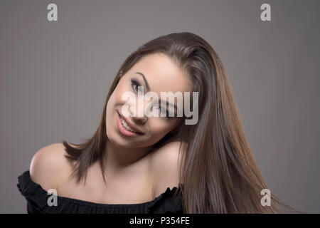 Charmante nette junge weibliche Mode Modell mit gesunden fließendes Haar lächelnd und mit Blick auf die Kamera auf grauem Hintergrund. Stockfoto