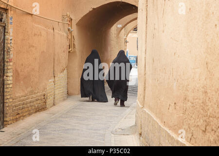 Yazd, Iran - 23. März 2018: Zwei muslimischen Frauen, im schwarzen Tschador Spaziergang auf der schmalen Straße der alten Stadt Yazd gekleidet Adobe Stockfoto