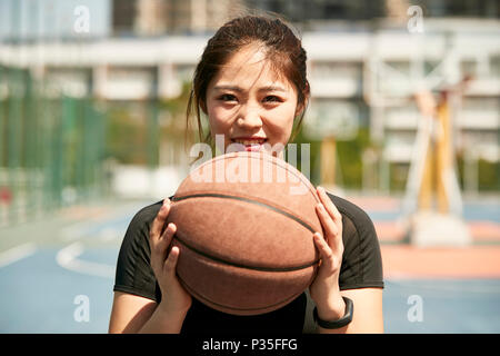 Junge asiatische Frau mit einem Basketball an der Kamera schaut lächelnd. Stockfoto