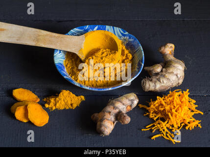 Kurkuma Wurzel und geriebenen frischen Kurkuma mit Pulver und hölzerne Schaufel auf rustikalen schwarz Tabelle Stockfoto