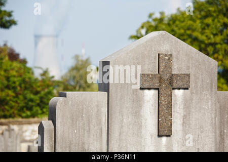 Ein Grabstein mit einem Kreuz auf einem Friedhof, ein Kernkraftwerk in den Hintergrund. In Huy, Tihange, Belgien. Stockfoto