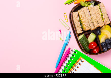 Overhead schiessen mit Holz- Lunch Box mit Sandwiches, Früchte und Gemüse auf rosa Hintergrund und Schule Schreibwaren. Gesunde Kinder Konzept essen. Stockfoto