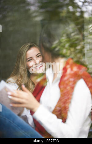 Hübsches Mädchen lächelnd zu ihr Freund hinter Fensterglas sitzen. Die Frauen, die klassischen, weißen Hemden und Strickjacken, blaue Jeans. Kaffee trinken, eine gute Zeit habend, die ror-Projekt. Stockfoto