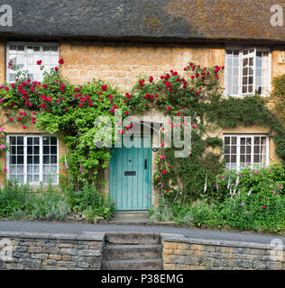 Reetdachhaus und rote Rosen um die vordere Tür in Ebrington, Chipping Campden, Gloucestershire, England klettern Stockfoto